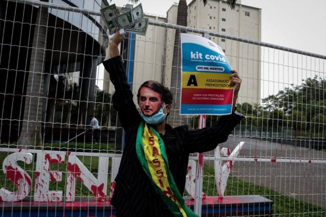 A imagem mostra uma pessoa com máscara de Jair Bolsonaro e faixa presidencial; em uma das mãos ela carrega um cartaz que simula a caixa de um medicamente que fez parte do chamado kit covid e na outra papeis que simbolizam dolares