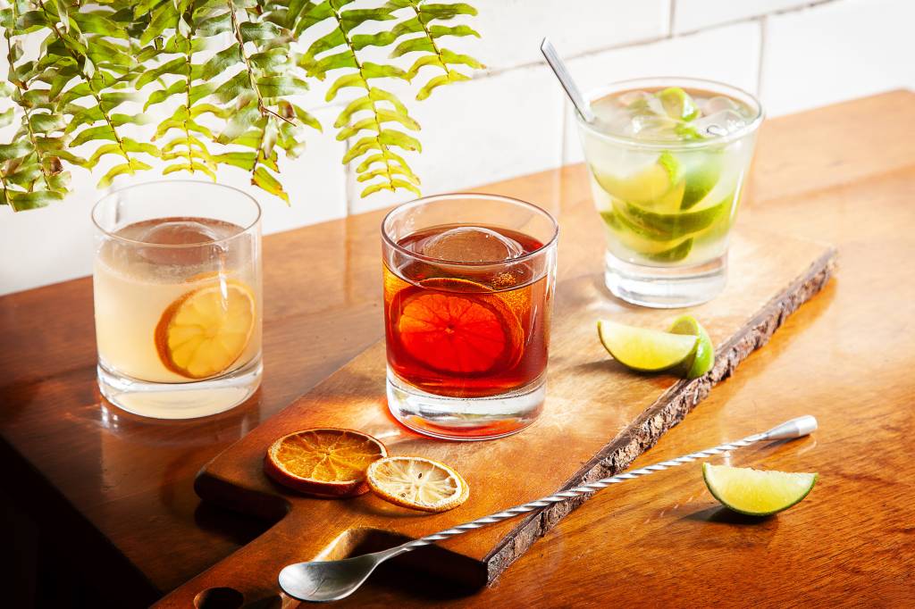Achados ELO – Samambaia Bar e Lanches Três copos de drinques, um boulevardier, uma caipirinha e um fitzgerald sobre tábua em cima de mesa de madeira.