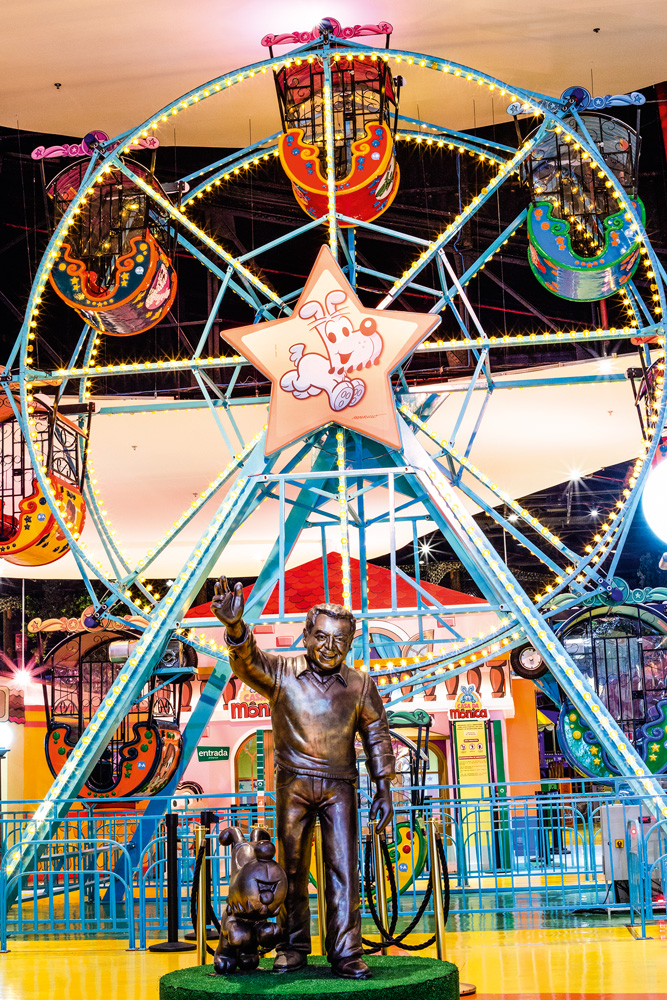 Nova Roda Gigante (5).jpg Uma foto de uma roda-gigante estampada com vários personagens da Turma da Mônica. Na frente do brinquedo, uma estátua em bronze de Mauricio de Sousa
