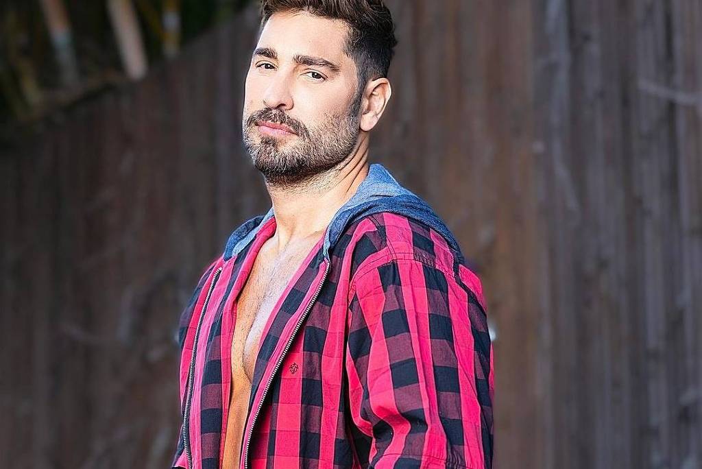 Victor Pecoraro posa com camisa quadriculada nas cores vermelho e preto, olha para a câmera sem sorrir. Tem barba e cabelo curto.