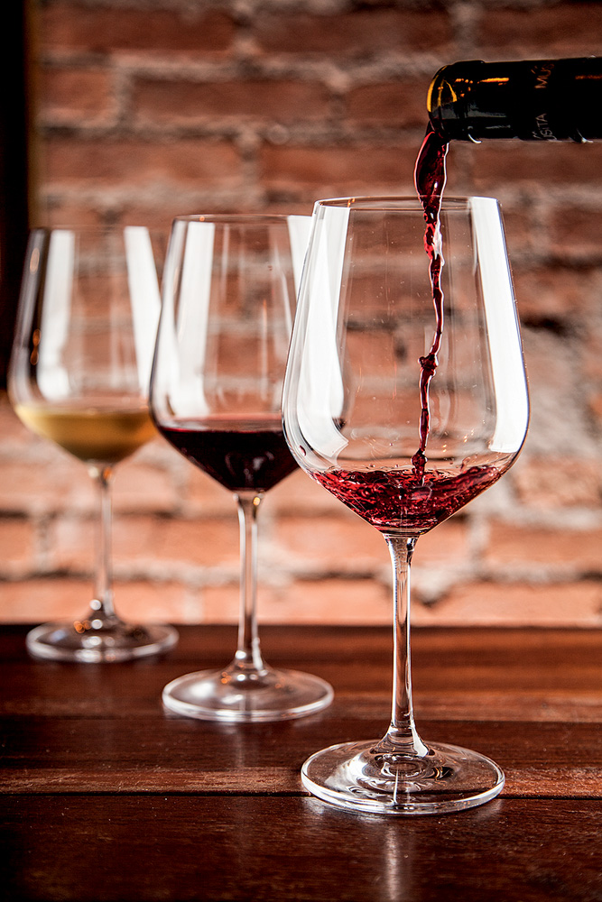 Sede261 Três taças de vinho alinhadas em diagonal sobre mesa de madeira. Ao fundo com vinho branco, no meio com tinto e na ponta com tinto sendo servido.