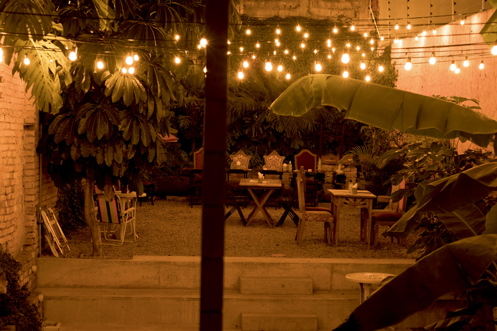 jardim-aberto-casa-cultural-Ala-Jardim vista noturna, com luzes amarelas dispostas em fios suspensos, do jardim aberto do espaço, com grama, plantas, mesas e cadeiras