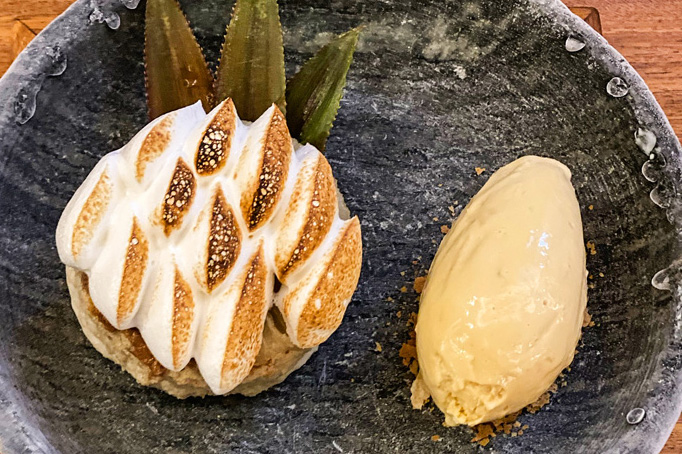 Sobre um prato fundo de cor escura, um bolo esponja de rapadura com abacaxi confitado e coberto por merengue maçaricado. Ao lado, o sorvete feito à base de shoyu sobre farofa doce crocante.