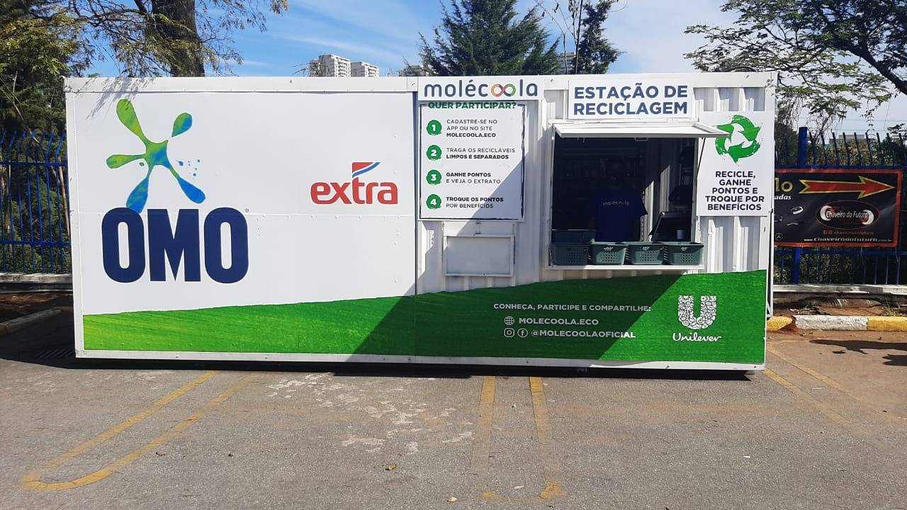 container de reciclagem branco e verde com logos da omo e do extra, posicionado no estacionamento do mercado