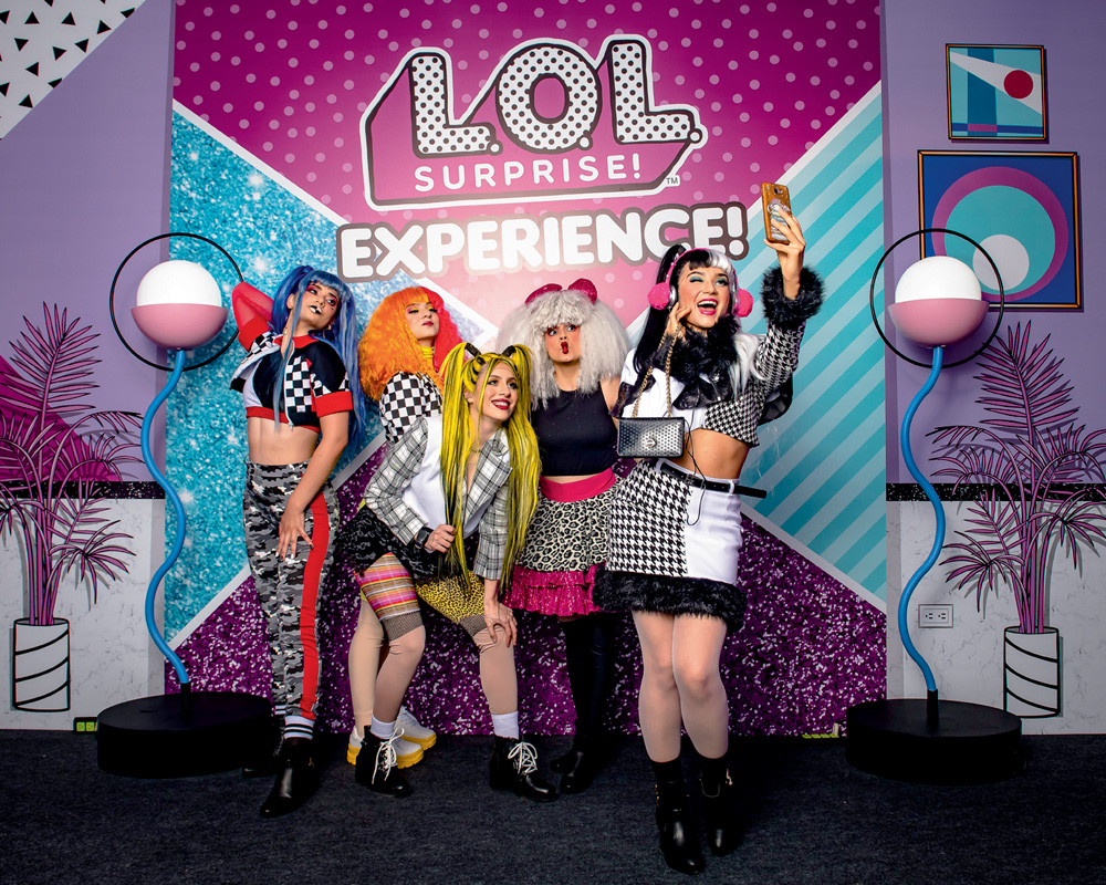 L.O.L. Surprise! Experience Em um cenário rosa, azul e roxo, cinco mulheres vestidas de bonecas L.O.L. posam para a foto. Elas vestem perucas e roupas chamativas e coloridas. Atrás, um banner escrito