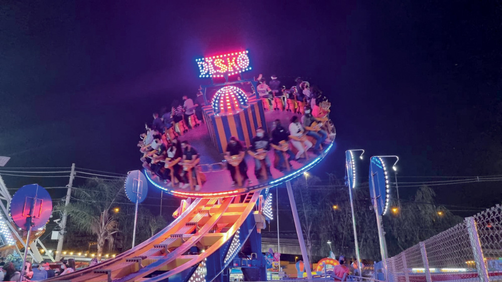 À noite, um brinquedo de parque de diversões diverte as pessoas. Ele é todo iluminado com LEDs roxas e rosas. O brinquedo é como um disco que gira em torno de si mesmo e também em uma estrutura presa ao chão