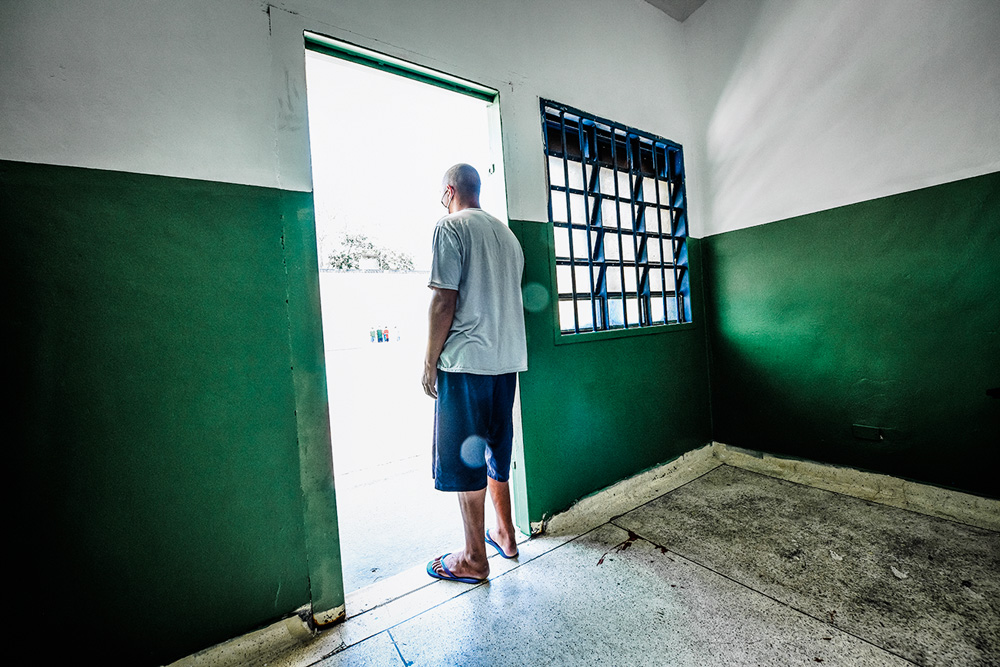 FUNDCASA_VIDANOVA_7.JPG A imagem mostra um detendo, apoiado em uma porta que dá à um ambiente externo, com a luz do sol bem forte.