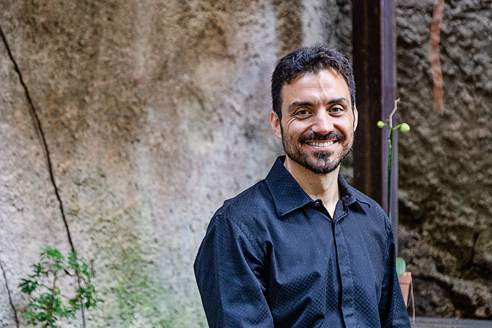 Matheus Macêdo Um homem de camisa social preta posa sorrindo para a foto, no que parece ser um quintal