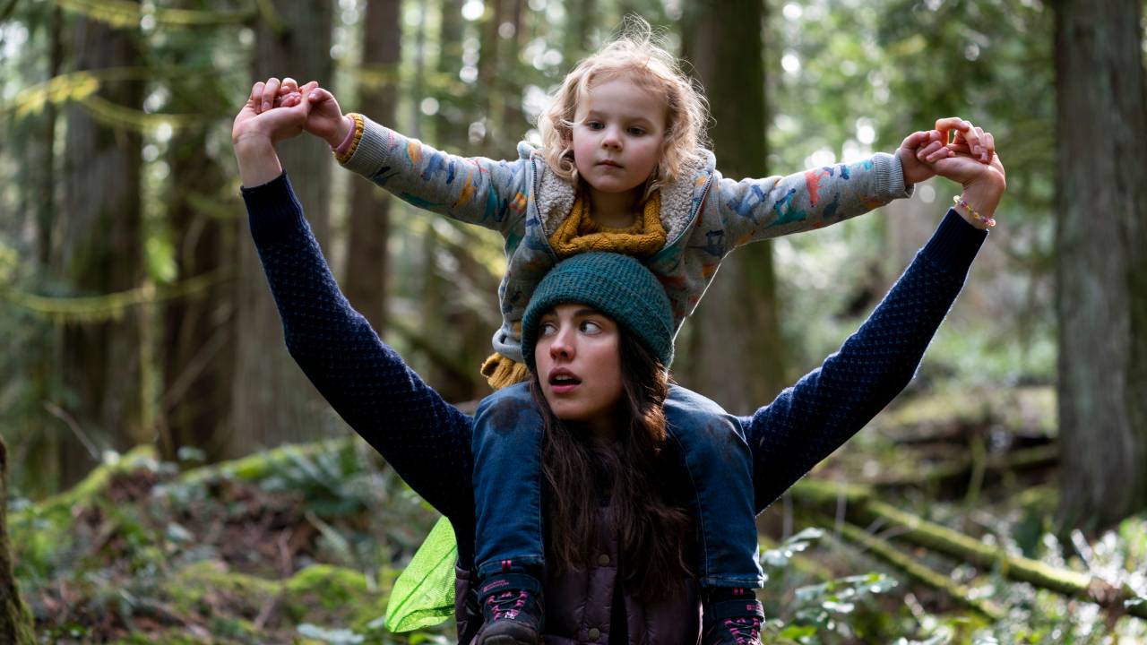 A imagem mostra uma jovem com sua filha em seus ombros, em uma floresta