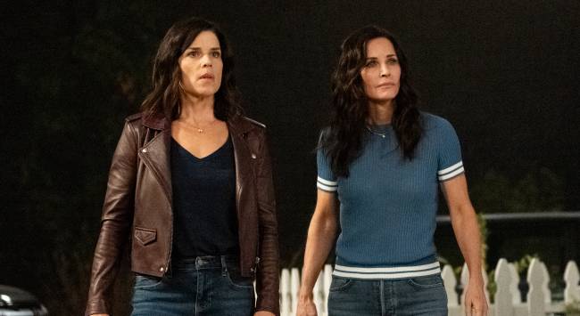 A imagem mostra as atrizes Neve Campbell (Sidney Prescott) e Courteney Cox (Gale Weathers), estrelas de Pânico, olhando assustadas para algo