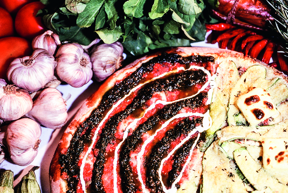 No canto superior esquerdo est&atilde;o cabe&ccedil;as de alho ao lado de folhas frescas de manjeric&atilde;o. Na por&ccedil;&atilde;o central, meia pizza com metade de abobrinha e metade de molho de tomate com pesto.