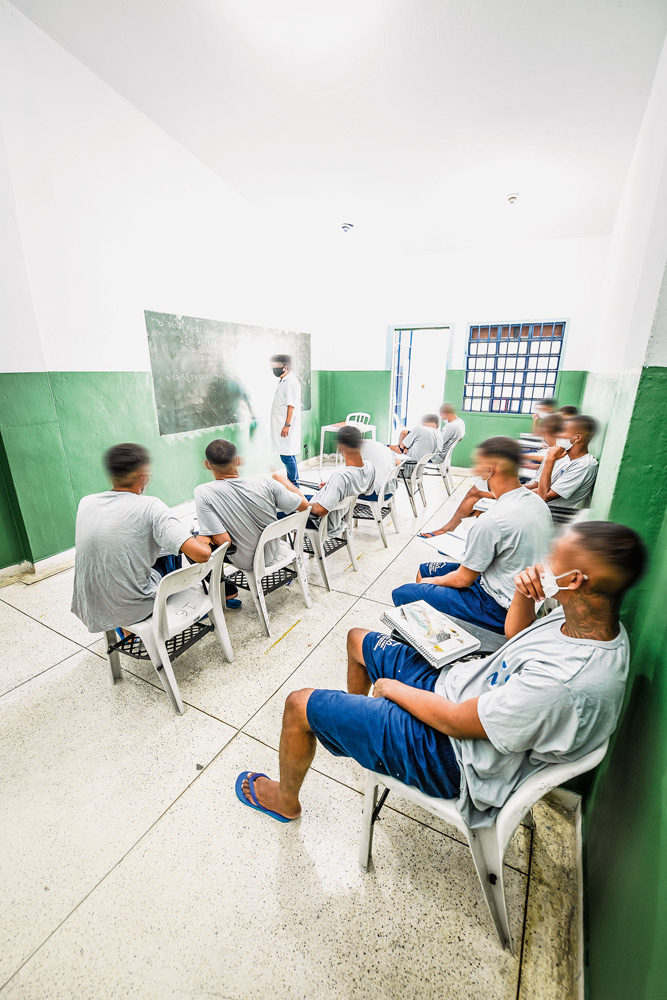 fundação casa sala de aula A imagem mostra alunos sentados em cadeiras de plásticos, vendo um professor na frente da sala, escrevendo em uma lousa.
