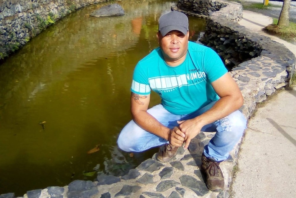 orazil agachado posando para a fota de boné, camisa azul e jeans próximo a um lago