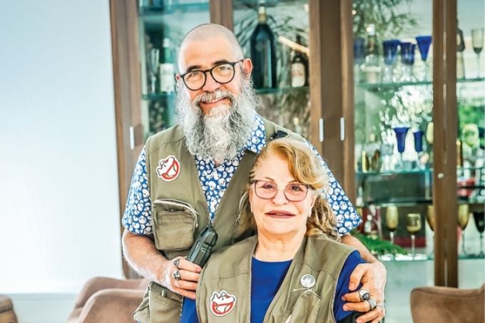 rosa maria e joão com jalecos de caça-fantasmas posando para a foto; eles estão sorrindo e sentados em uma poltrona, ela no assento e ele no braço do móvel