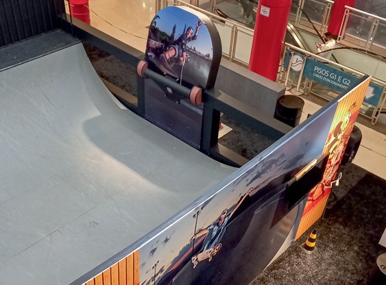 Dentro de um shopping tem uma rampa de skate branca dentro de um espaço fechado. A foto está de cima e há banners e placas de skate para decorar