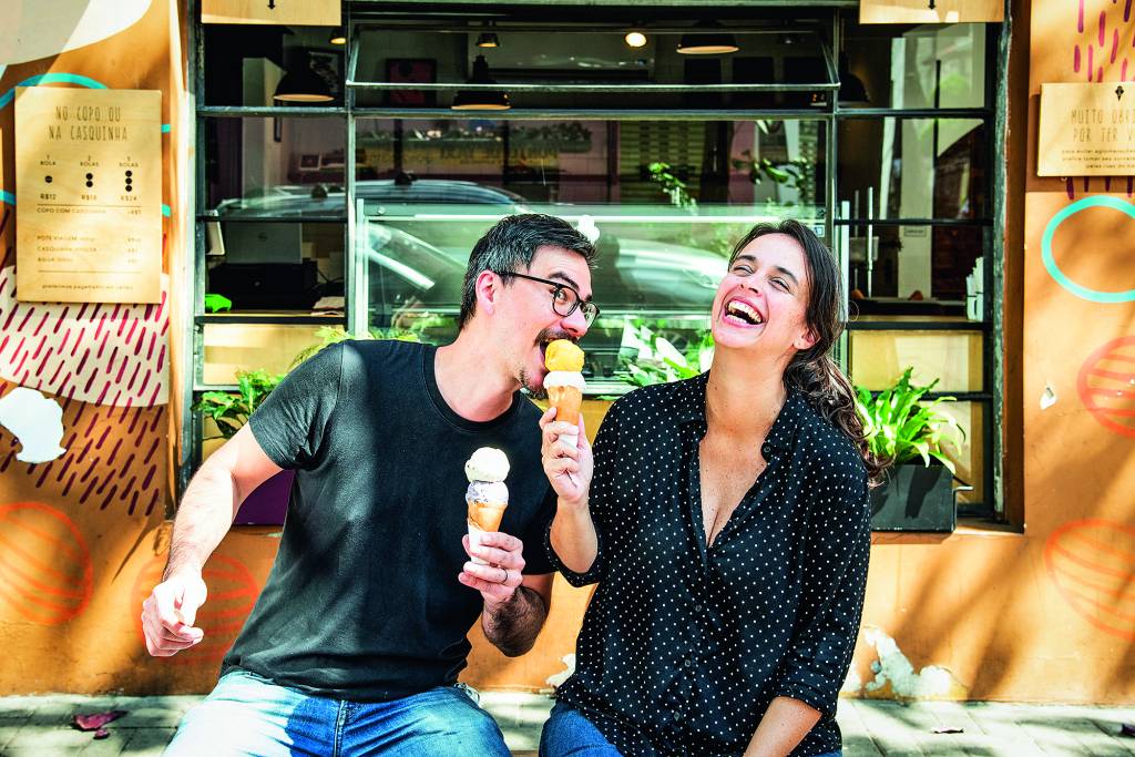 COMER & BEBER 2021 – FRIDA E MINA O casal Thomas Zander e Fernanda Bastos sentados em frente à Frida & Mina com ele tomando da casquinha de sorvete dela enquanto ela sorri.