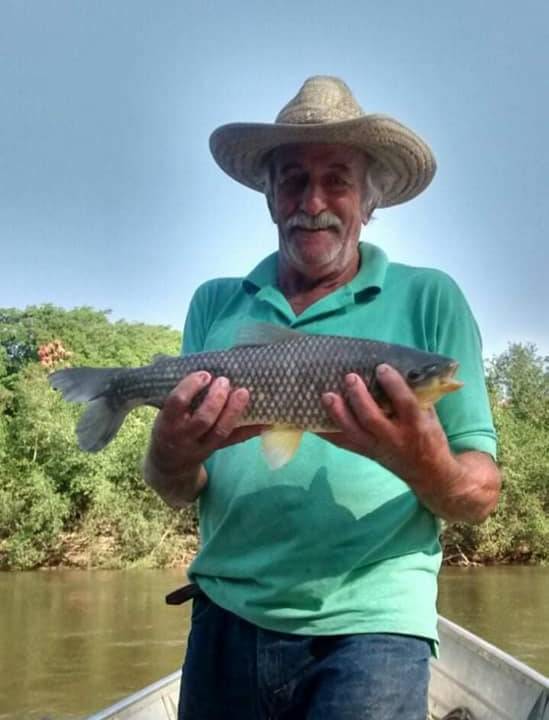 Imagem mostra homem de chapéu de palha segurando peixe em barco, no meio de rio