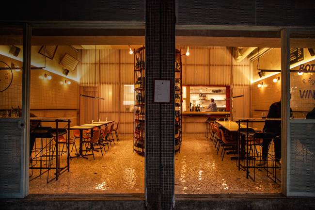 Ambiente do bar de vinhos Paloma, cujas paredes s&atilde;o forradas por azulejos de cor branca. Garrafas de vinho expostas pelo sal&atilde;o tamb&eacute;m fazem parte da decora&ccedil;&atilde;o.