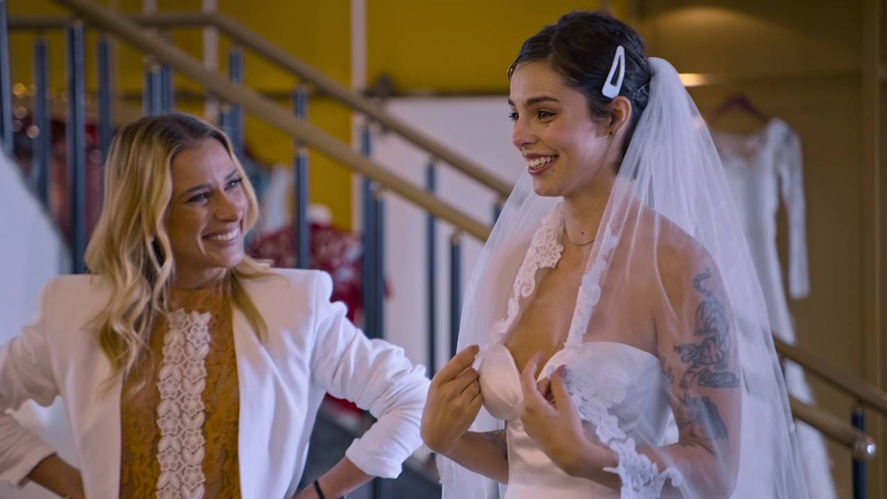 Lethicia sorri ao olhar noiva, que encara emocionada o vestido no espelho.