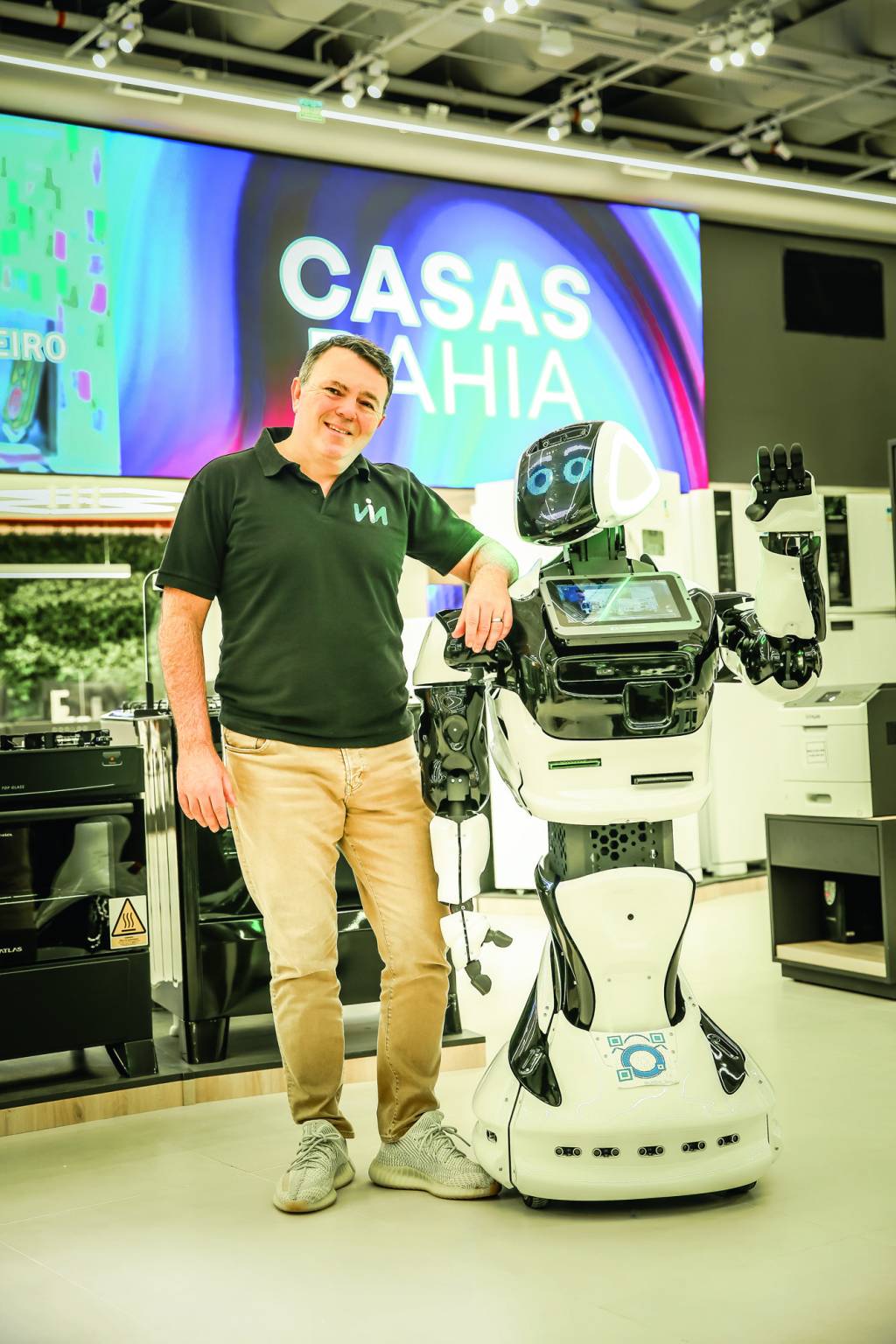 CASASBAHIA_21.JPG Homem com calça marrom e camisa polo verde escura posa com o braço no ombro de um robô branco e espelhado.
