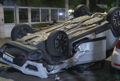 Imagem mostra carro branco capotado, com as rodas para cima no meio de uma rua.