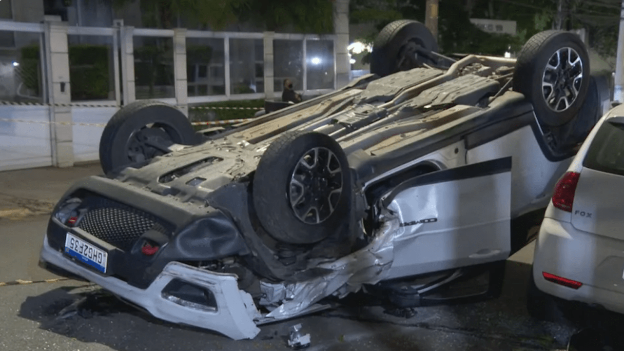 Imagem mostra carro branco capotado, com as rodas para cima no meio de uma rua.