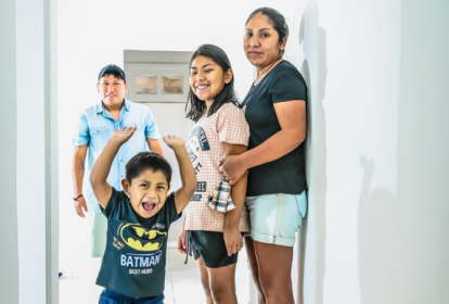 Imagem mostra uma família com um homem, uma mulher e dois filhos, uma menina e um menino. O menino levanta os braços e sorri, enquanto o pai está no fundo e a mãe abraça sua filha encostada na parede, no lado direito da imagem.