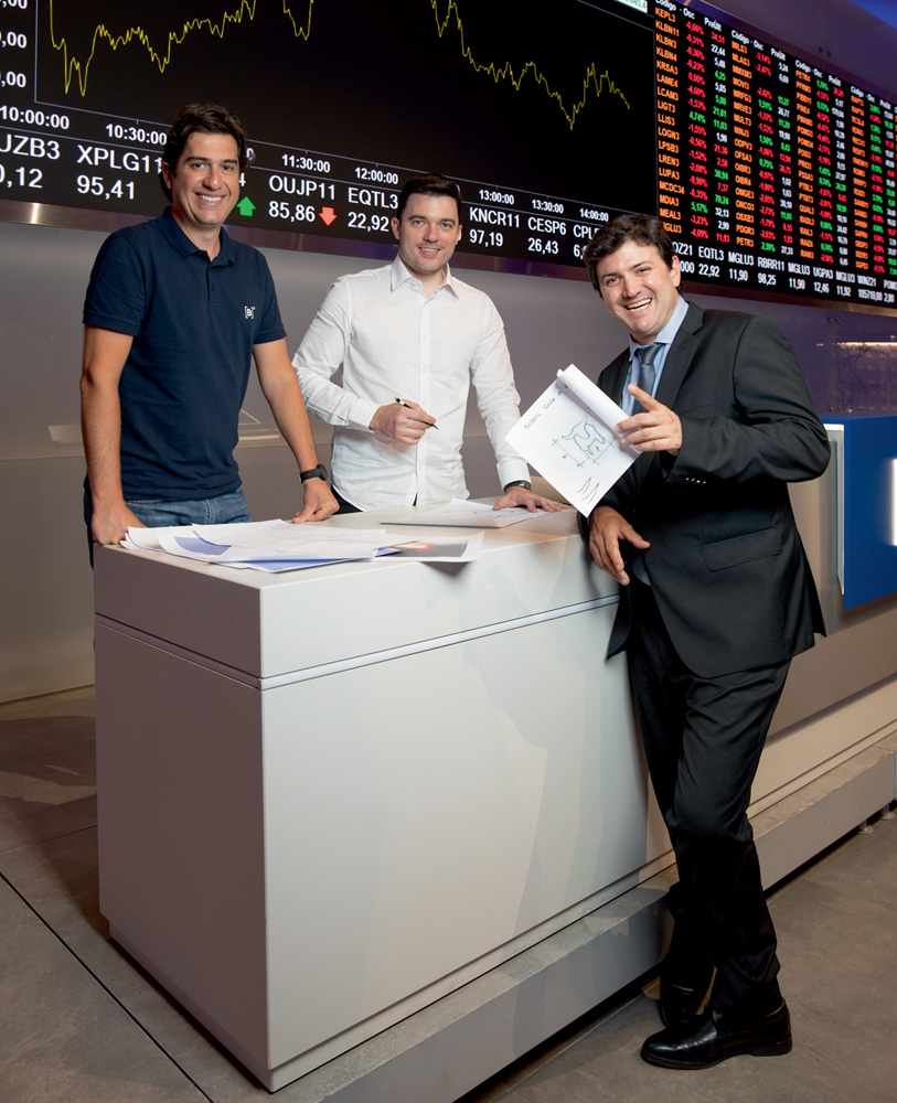 Touro da B3 A partir da esquerda, Felipe, Rafael e Pablo sorriem reunidos em um balcão branco. Ao fundo, painéis da bolsa de valores.