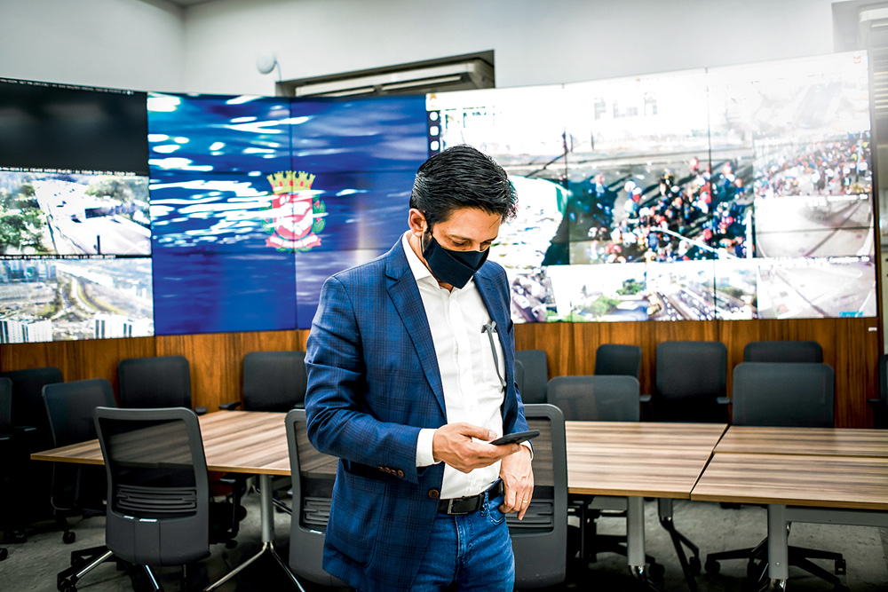 Ricardo Nunes O prefeito Ricardo Nunes está em uma sala com várias televisões que transmitem imagens da cidade. Ele está mexendo no celular