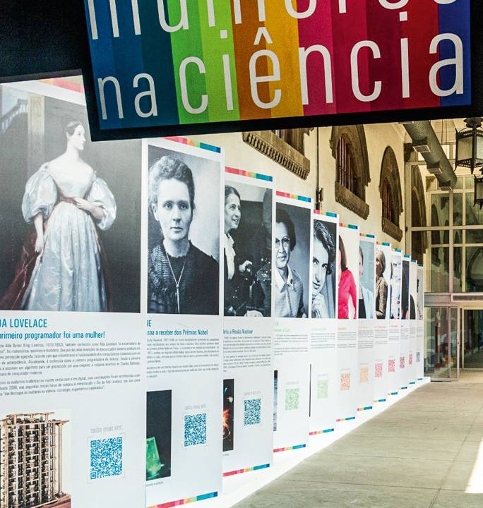 Em um corredor, estão enfileirados grandes banners com fotos e informações sobre diferentes mulheres cientistas. Há, no início do corredor, uma placa escrito "Mulheres na Ciência"