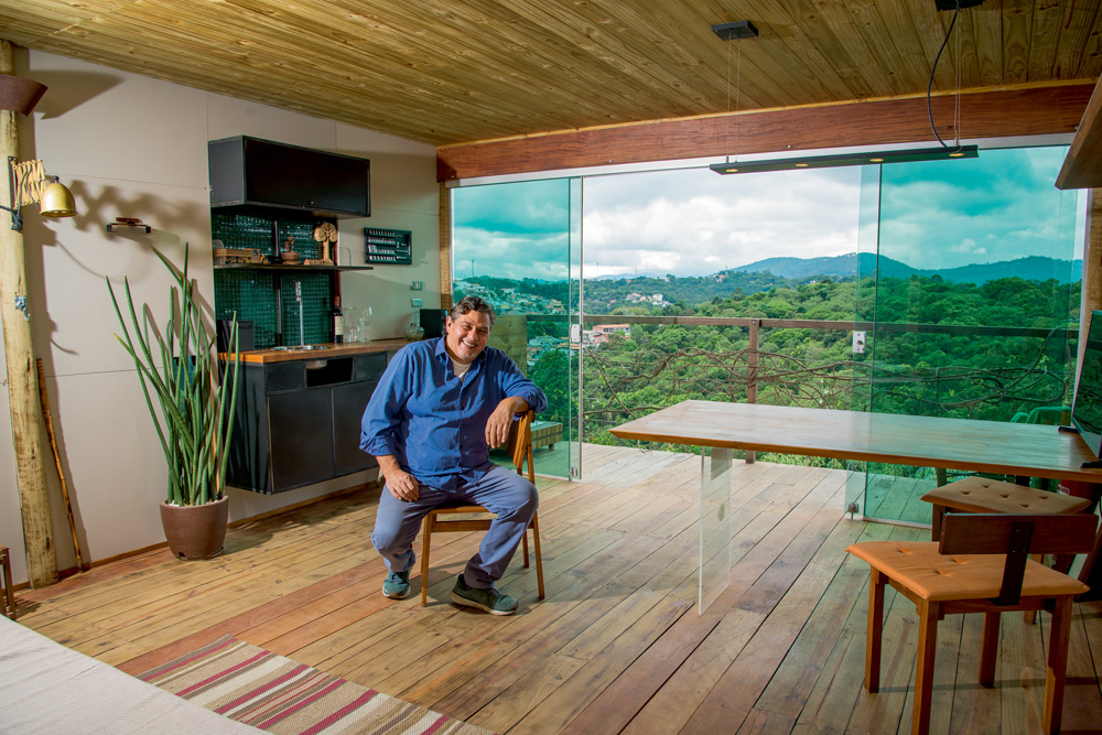 Veja SP A imagem mostra Ricardo Afiune sentado em um banco de madeira em uma sala repleta de móveis de madeira. Ao fundo, uma varanda que dá vista em um lugar cheio de árvores e verde.