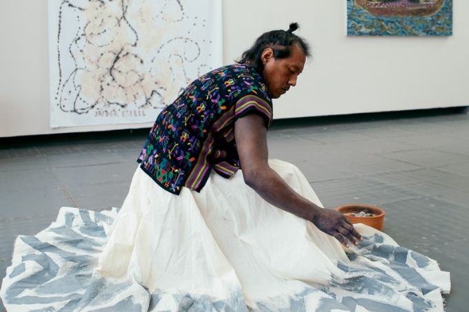 jaide esbell utilizando roupas indígenas em galeria de arte, de lado para a câmera, agachado sobre um pano sobre o qual ele está pintando
