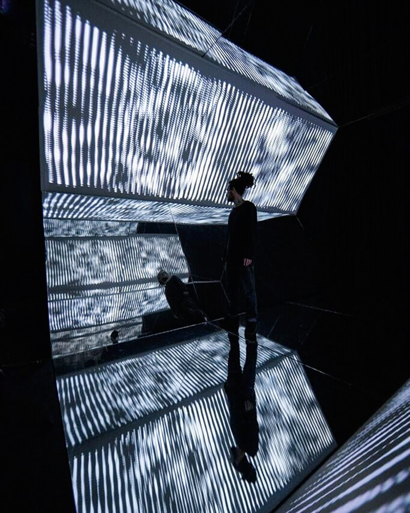 vj-vigas-aparato-10-farol-santander-obra-de-arte-luminosa-videomapping foto de VJ Vigas dentro de sua obra aparato 10, que tem múltiplas estruturas geométricas luminosas. ele está na penumbra
