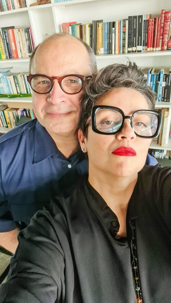 20211129_101952.jpg Imagem mostra uma mulher de roupa preta, óculos quadrados e batom vermelho e um homem de camisa azul detrás. Ao fundo, uma biblioteca.