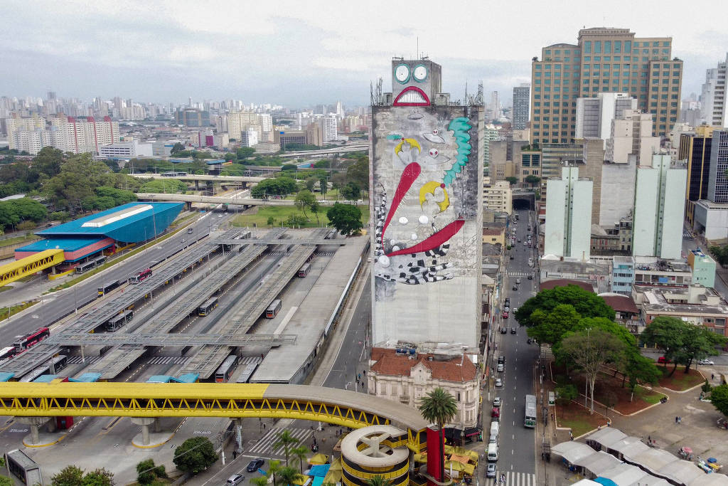 Na foto é possível ver uma imagem panorâmica da ilustração no edifício-garagem Parque 25, no centro de São Paulo. A obra cobre todo o paredão lateral do prédio e mostra um robô cinza sendo engolido por um monstro de lábios vermelhos e dentes pontiagudos, enquanto discos voadores flutuam próximos ao robô.