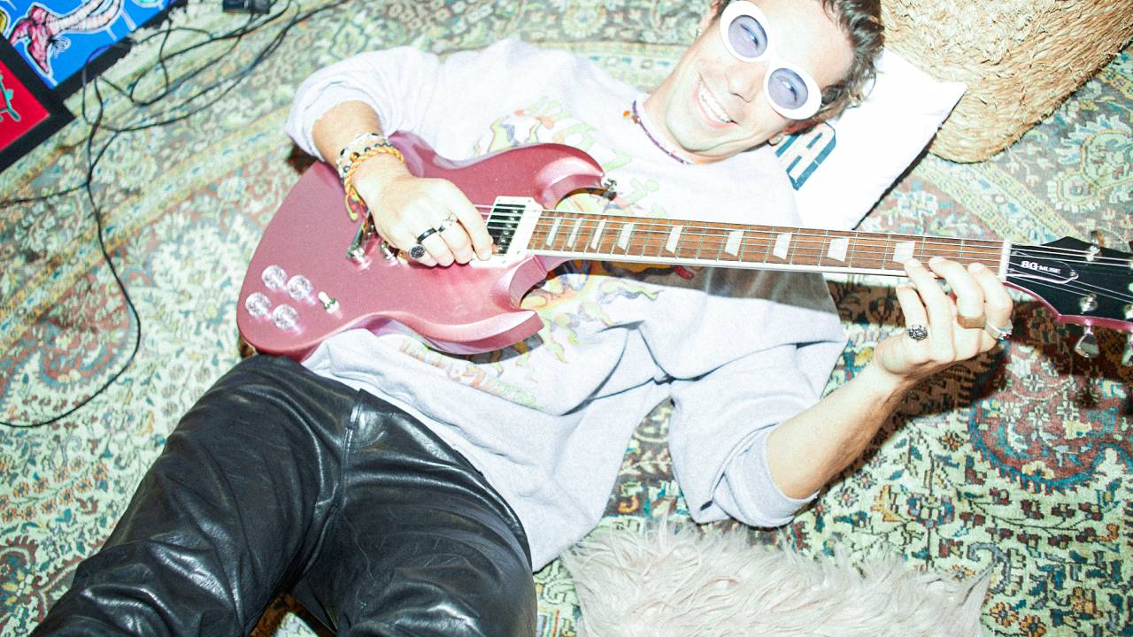 Imagem mostra homem deitado, sorrindo e de óculos escuros segurando uma guitarra vermelha.