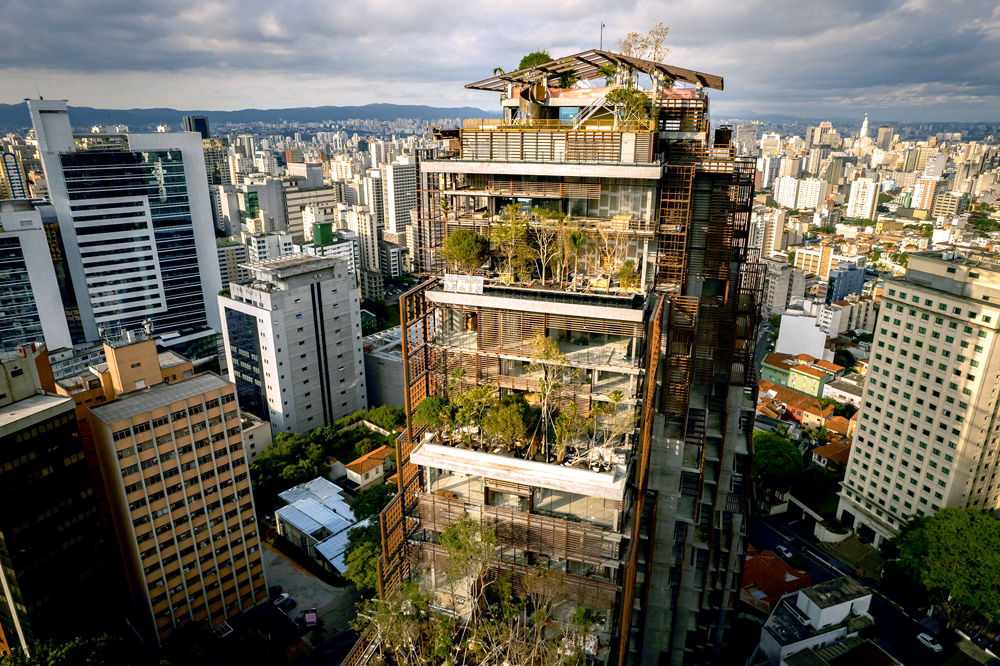 @gabbavisuals-0326.jpg Foto aérea mostra um edifício que tem seus andares todos arborizados e com muitas plantas. Dá para ver outras dezenas de prédios ao fundo, além do céu
