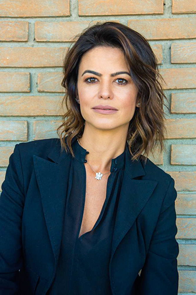 Adriana Drulla Adriana Dulla é uma mulher branca, de olhos e cabelos castanhos. Ela pose em frente a uma parede de tijolos e traja uma camisa azul e um colar prateado.