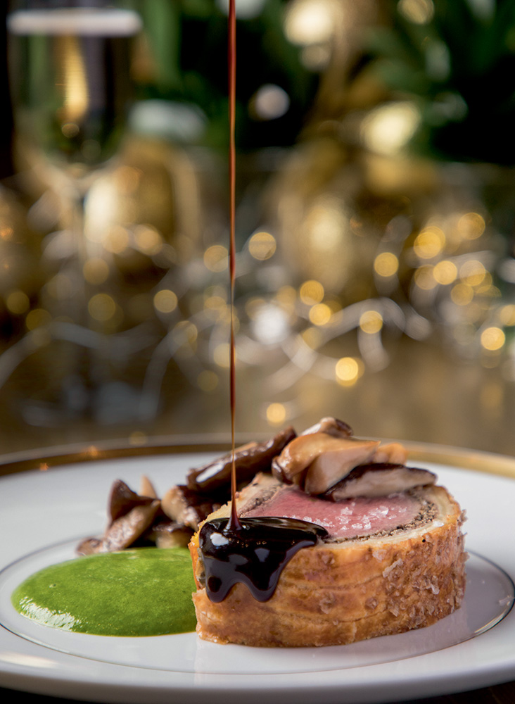 Tangará Jean-Georges Foto de fatia de bife wellington sendo coberto por molho escuro ao lado de creme verde de couve.