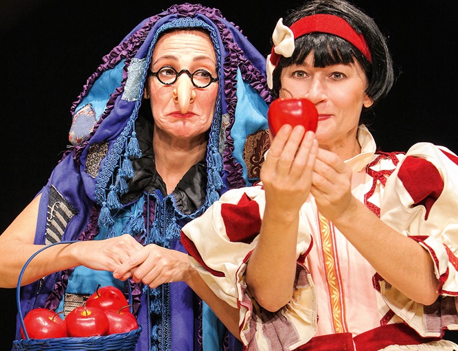 Duas mulheres estão em cena. Uma está vestida de bruxa má e a outra de Branca de Neve, que segura uma maçã