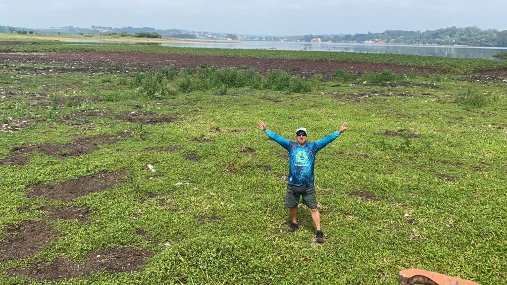 Projeto Limpeza na Represa Imagem mostra homem vestido de camiseta azul abrindo os braços em meio a gramado da represa de Guarapiranga