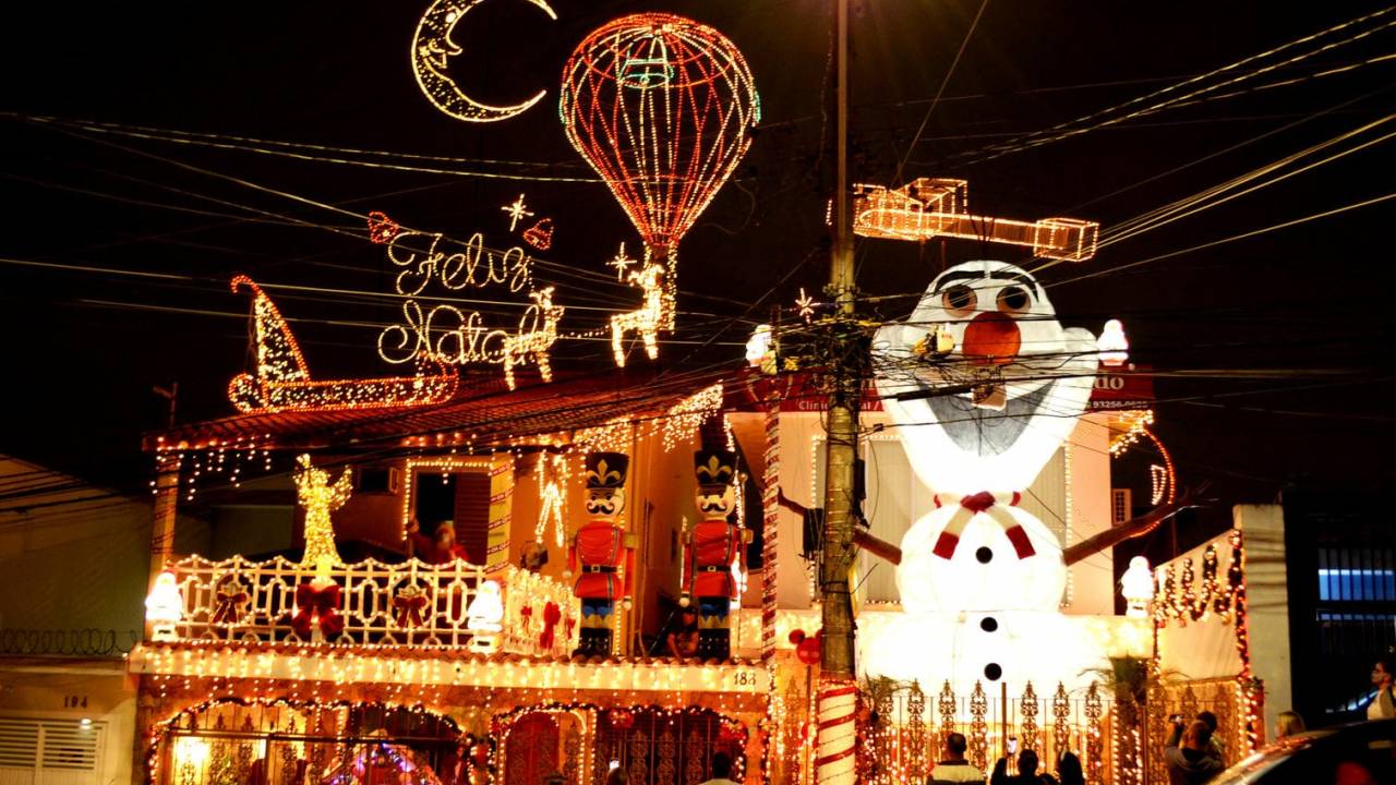 Uma residência de dois andares enfeitada com luzes e outras decorações natalinas. À esquerda, é possível ver um boneco gigante do Olaf, personagem do filme "Frozen". À direita estão soldadinhos, um trenó com renas todo iluminado e outros enfeites. As luzes vermelhas e amarelas cobrem toda a casa.