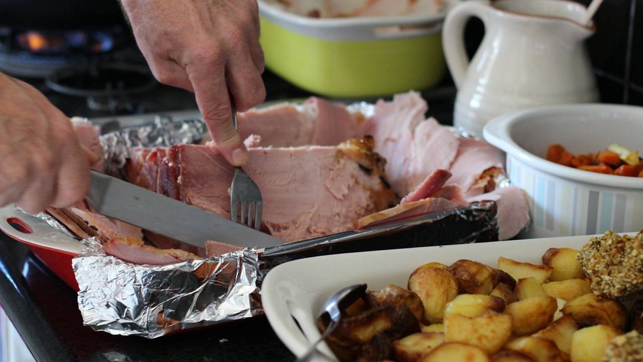 Imagem mostra mesa cheia de comida e mãos cortando pedaço de carne de porco com garfo e faca