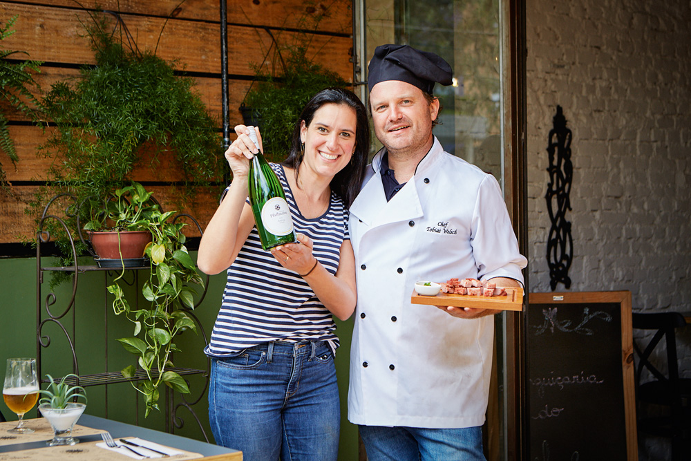 Imagem mostra um homem e uma mulher. Ele veste uma roupa de chef e ela levanta uma garrafa de espumante.