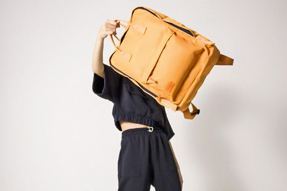 Mulher de roupa preta levanta no ar com uma m&atilde;o uma mochila quadrada e laranja.