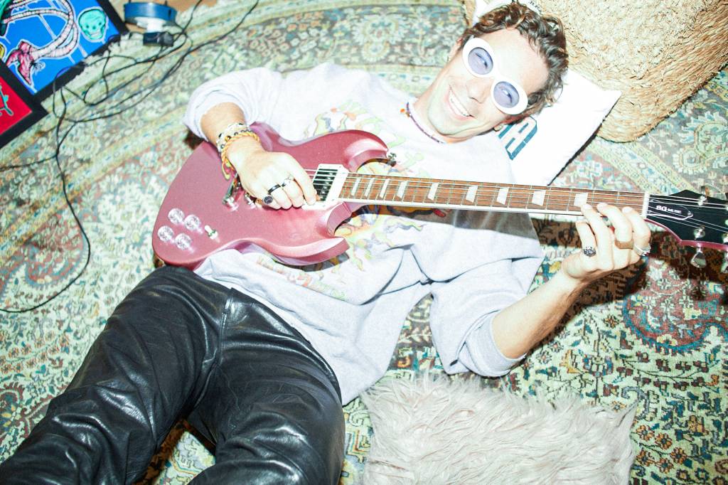 -61A9923–1- (1) Imagem mostra homem de calça preta e moletom branco deitado sobre tapete, sorrindo e tocando uma guitarra vermelha