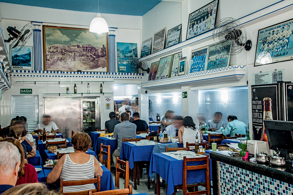 Ambiente do restaurante Acrópolis. Imagem mostra espaço de paredes brancas com mesas de toalhas azuis. Nas paredes, quadros também azuis e brancos.