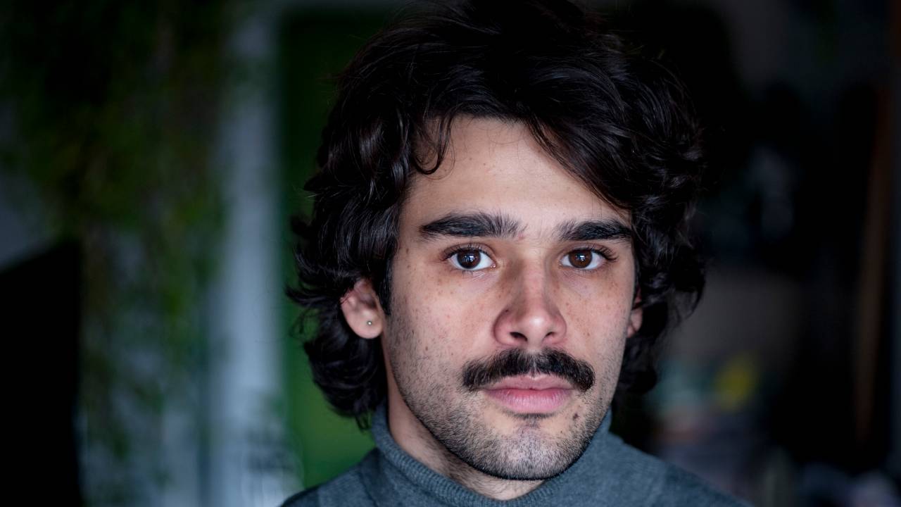 Imagem mostra homem de bigode e casaco cinza. Ele está olhando para a câmera.