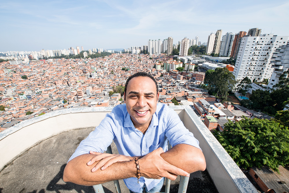 Créditos_Alexandre Battibugli.jpg (1) Imagem mostra homem de camisa azul e os braços cruzados. Ao fundo, uma favela.