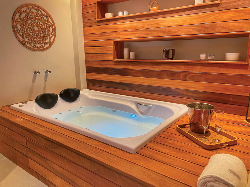 FOTO 9 – AURA SPA – foto divulgacao.jpeg Imagem mostra jacuzzi embutida em estrutura de madeira. Uma bandeja com duas taças está ao lado da jacuzzi.